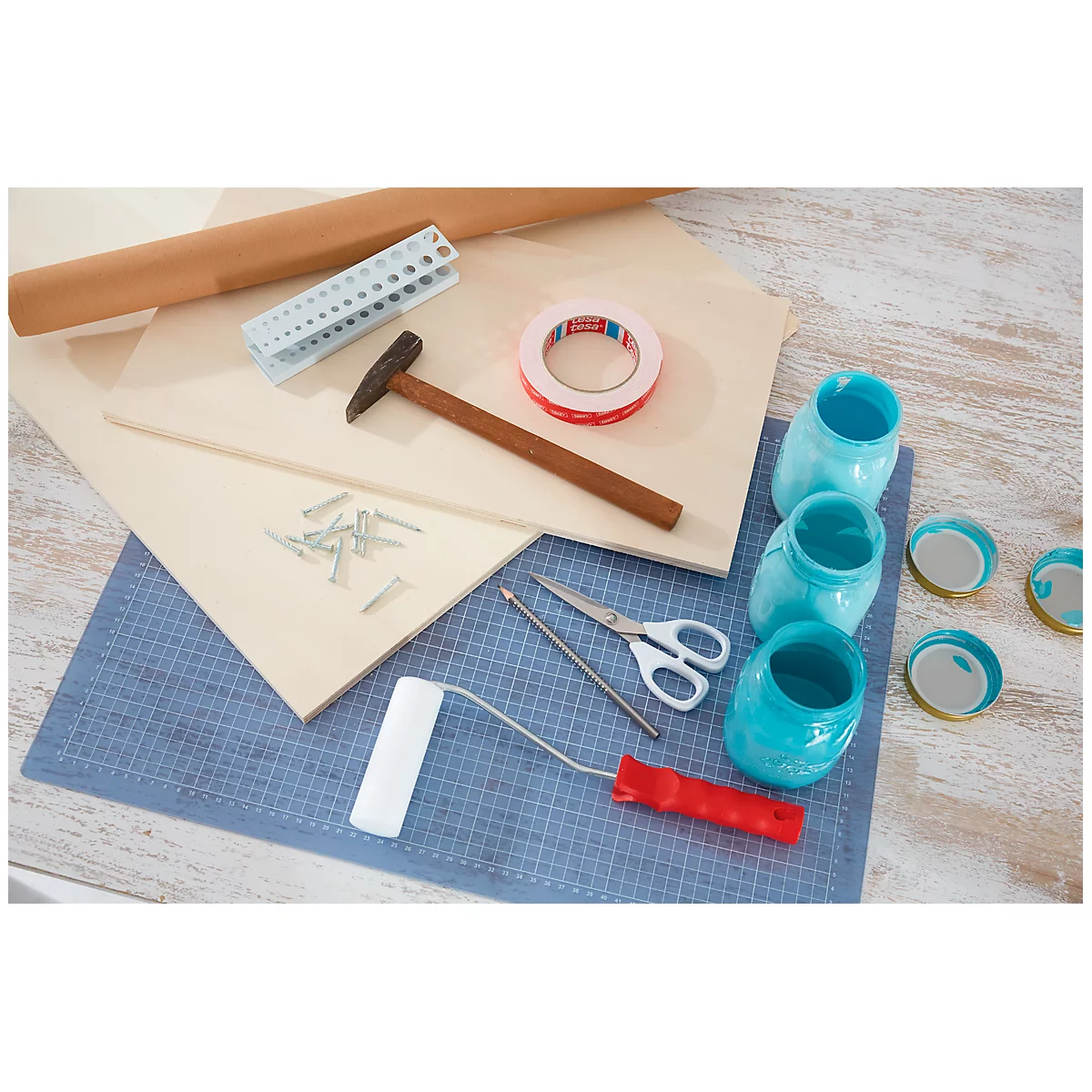 Close-up of tools and materials for DIY projects, including hammer, roller, paint jars and wood. All on a blue tablecloth.