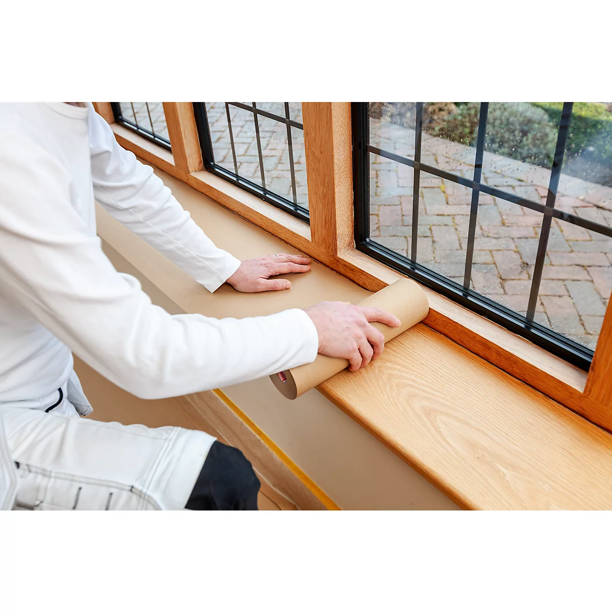 Person rollt Papier auf eine Fensterbank. Der Mann trägt weiße Kleidung und arbeitet an einem Holzfensterrahmen.