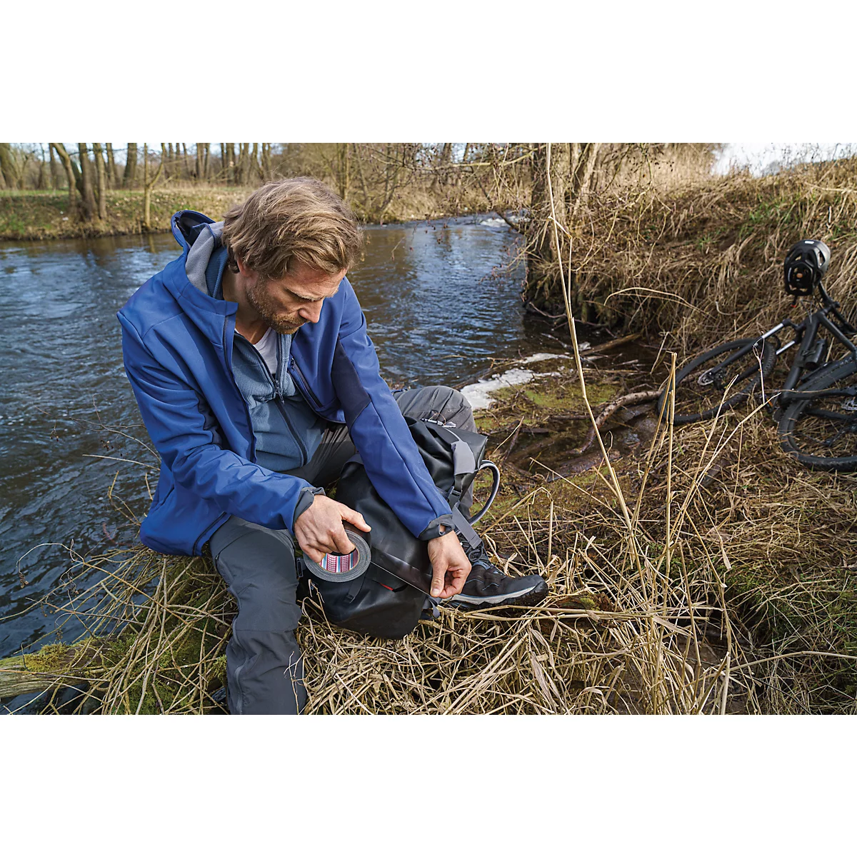 A man at the riverbank opens a black bag, a bicycle next to him. He wears a blue jacket.