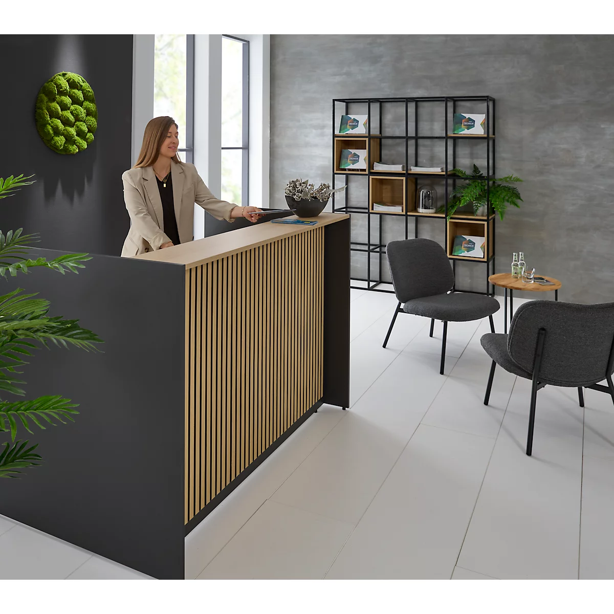 Eine Frau steht an einem Empfangstresen aus Holz und schwarzem Material. Im Hintergrund ist ein grünes Moosbild zu sehen.