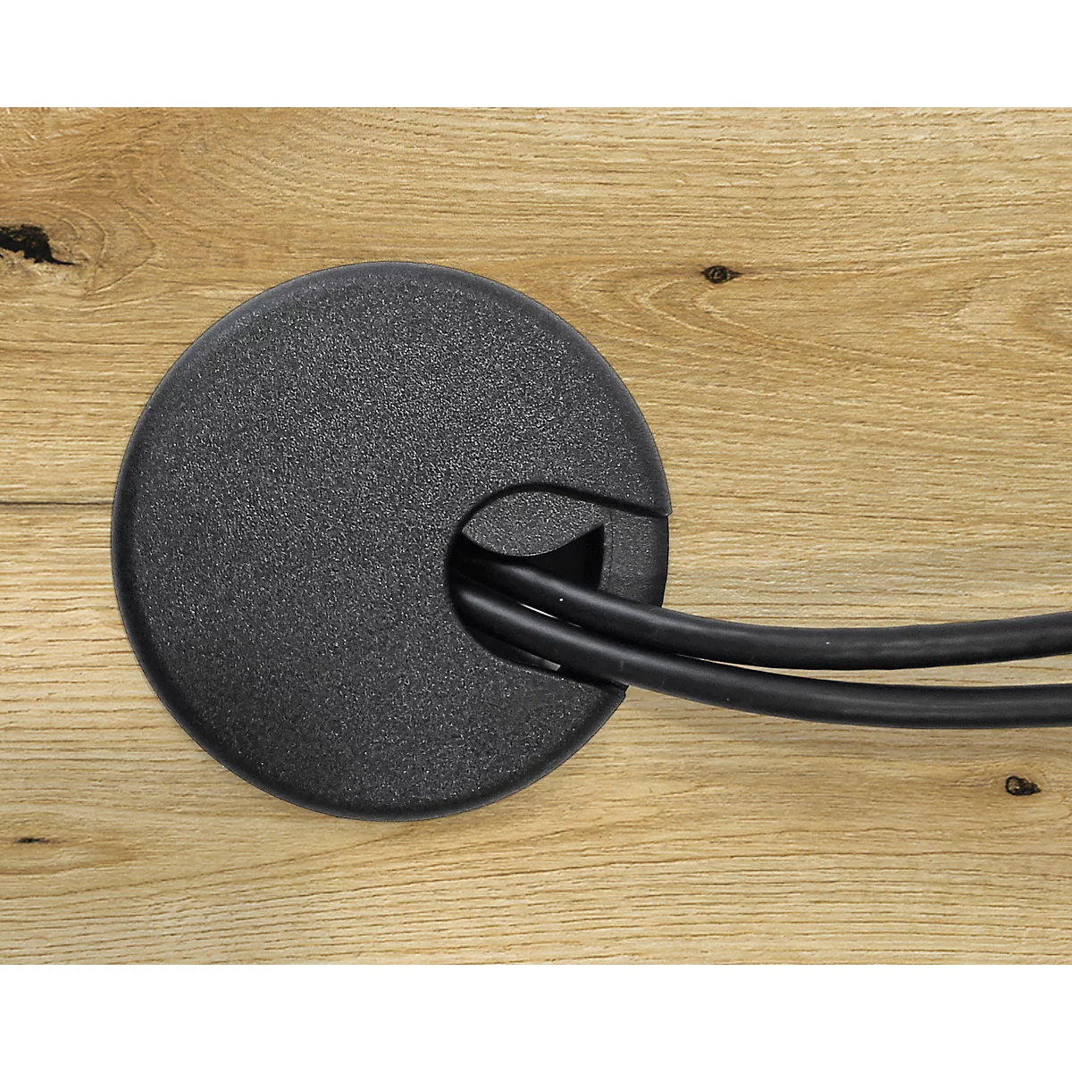 Schwarze Kabeldurchführung aus Kunststoff in Holzplatte. Zwei schwarze Kabel kommen aus der Öffnung.