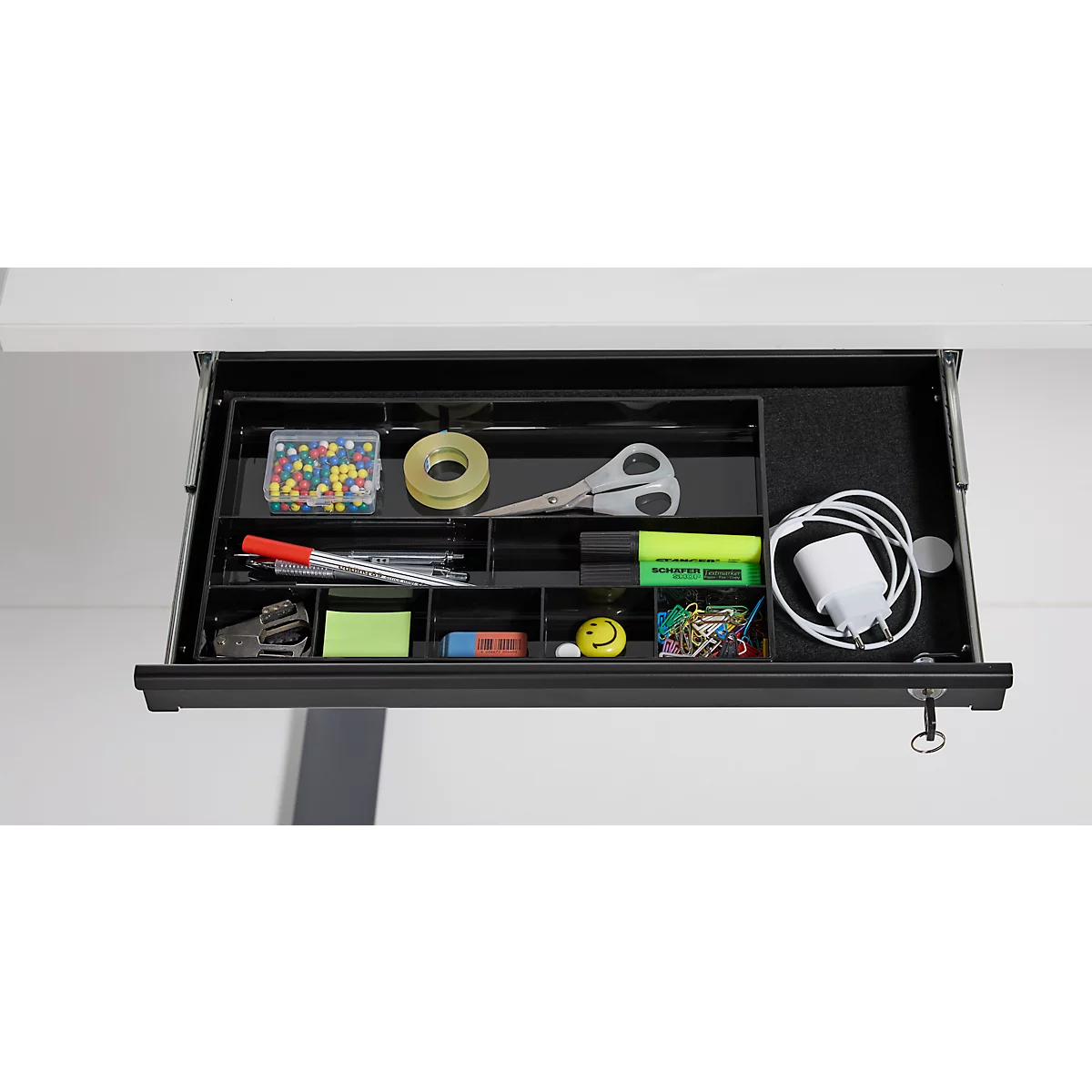 Tiroir noir ouvert avec fournitures de bureau: stylos, ciseaux, ruban adhésif, punaises, câble de chargement, marqueur et notes autocollantes.