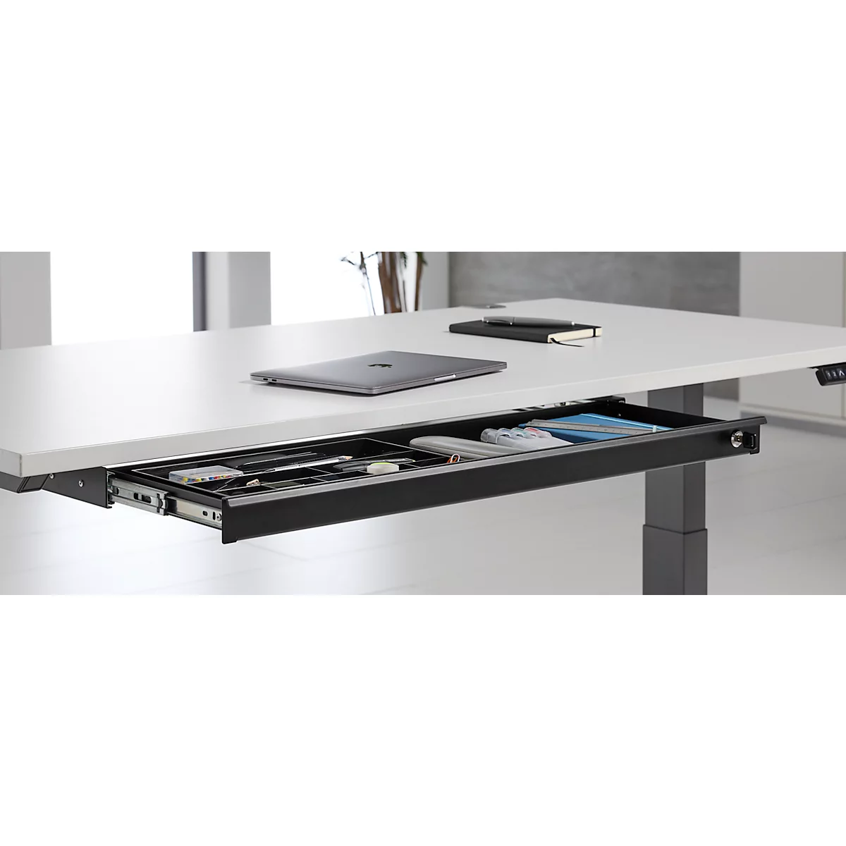 Bureau avec tiroir intégré, ordinateur portable, bloc-notes et stylo. Le tiroir contient des fournitures de bureau. Pieds de bureau gris.