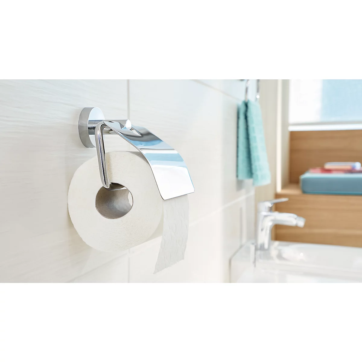 Chrome toilet paper holder with roll of paper; towel and sink in the background.