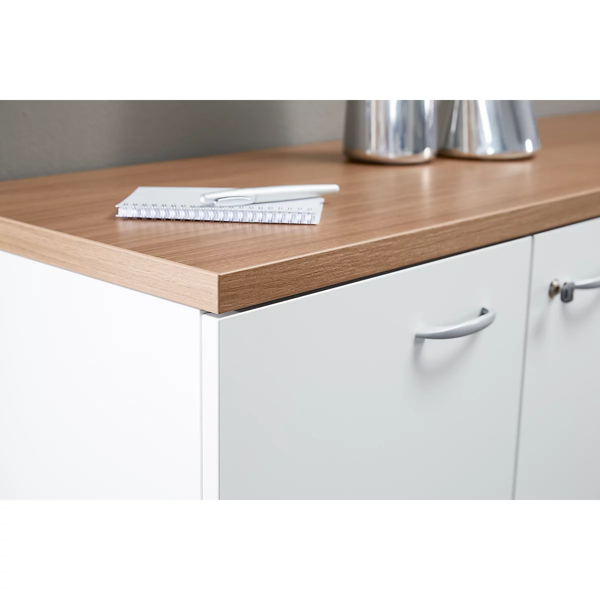 Close-up of white sideboard with wooden surface, on it notepad, pen and metal vases.