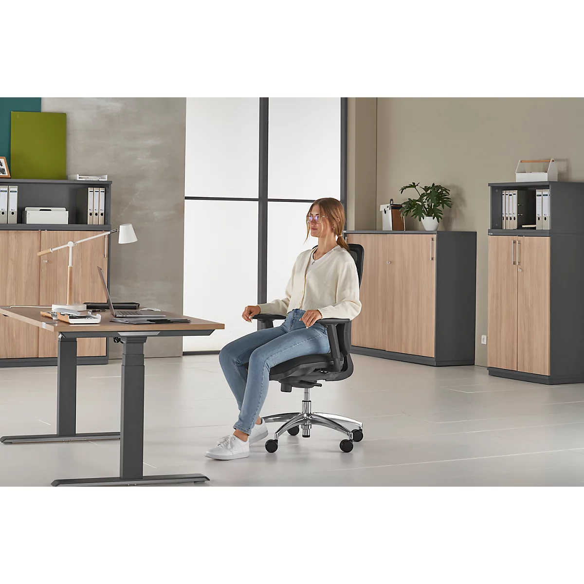 A woman sits in an office chair in front of a desk in an office. She is wearing jeans and a white sweater. The room is bright and modern.