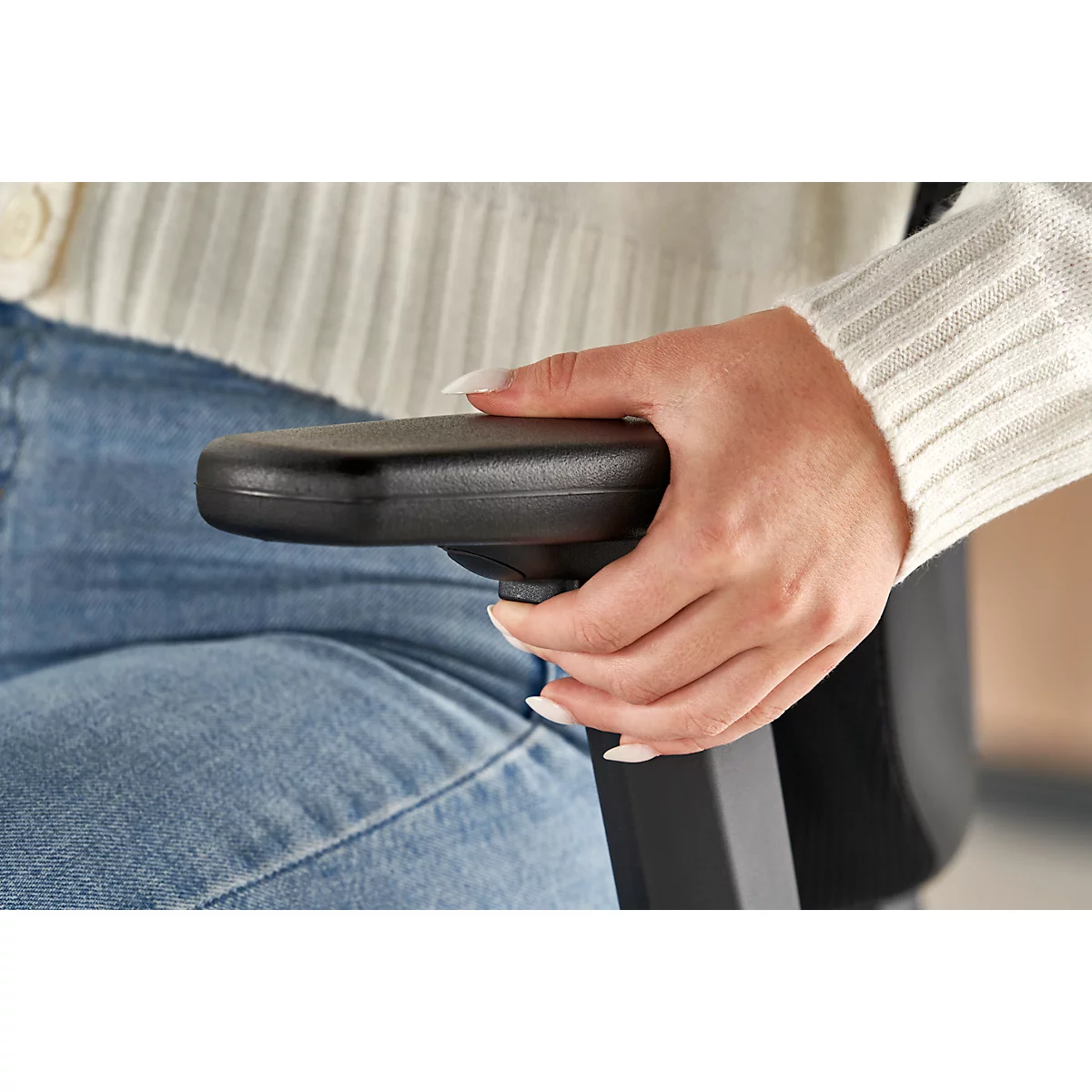 Close-up of a hand touching the armrest of a black office chair. The person is wearing jeans and a white sweater.