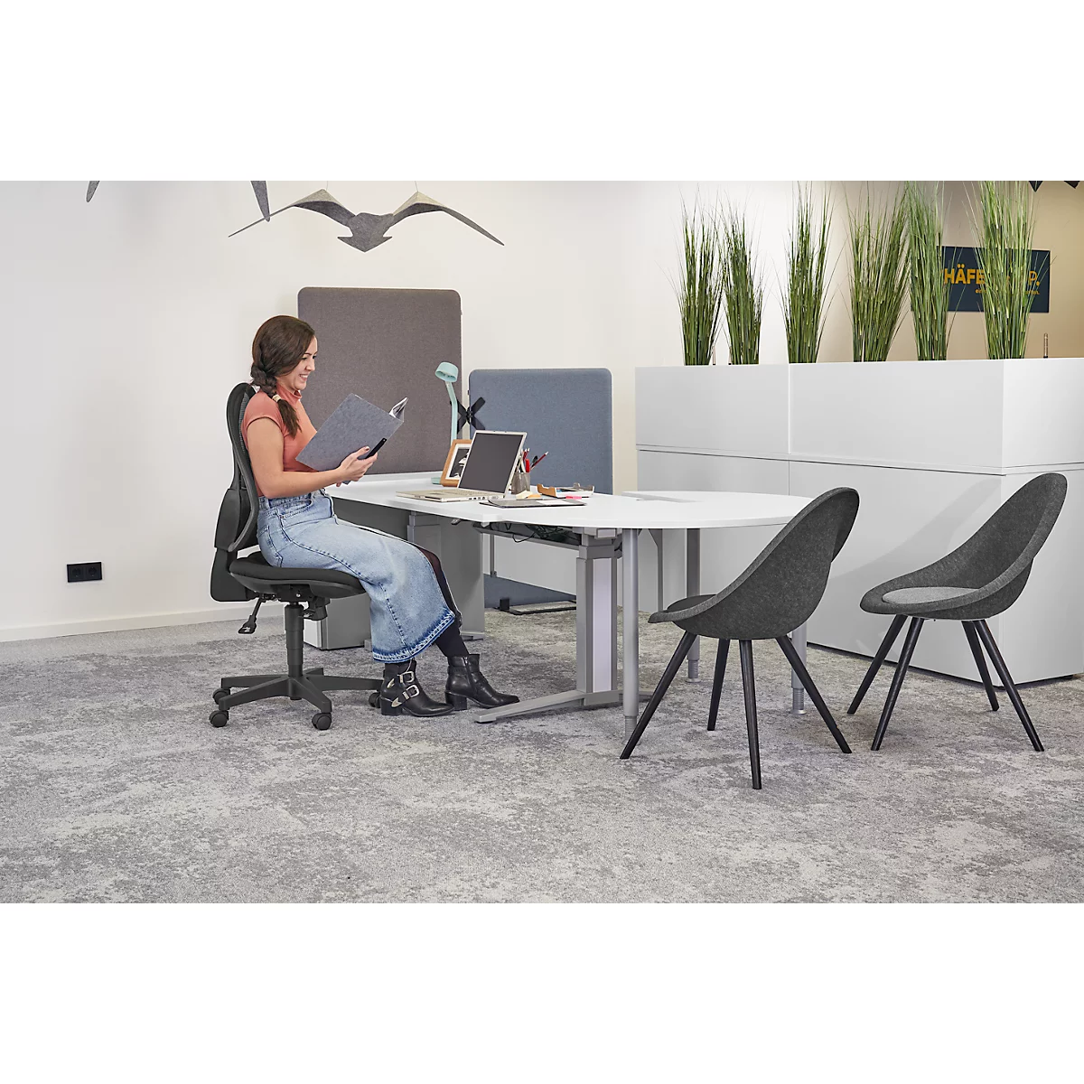 A woman sits at a desk reading, with office chairs and plants in the background.