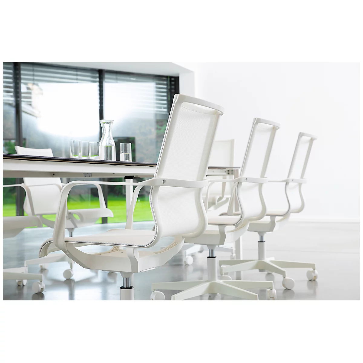 White office chairs around a table with glasses and a carafe, bright room, large windows.