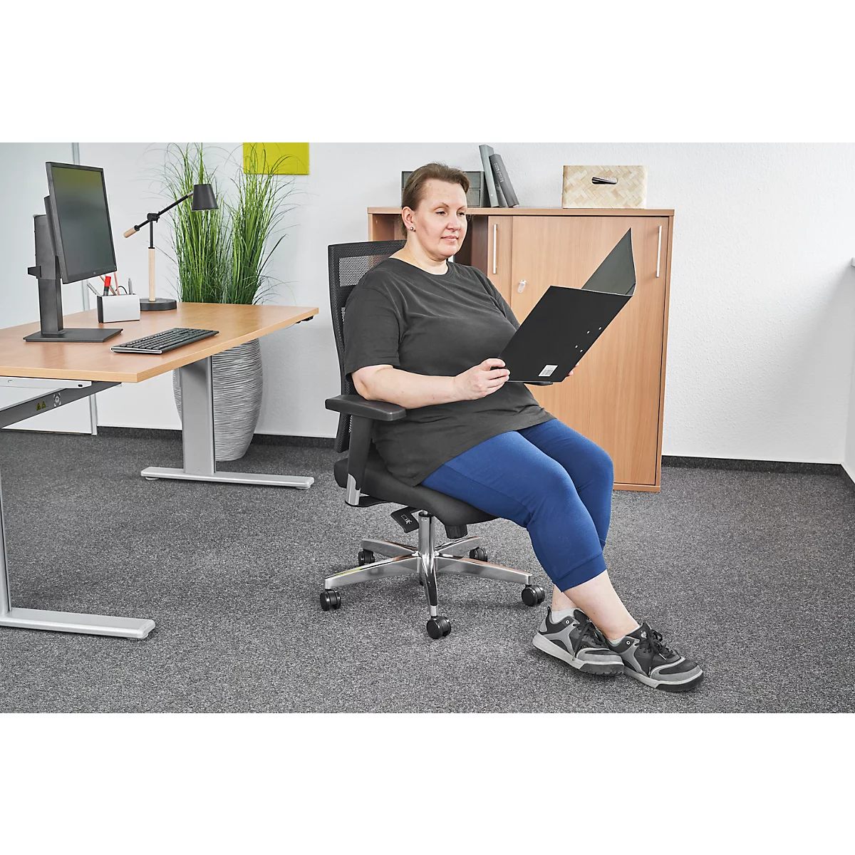 Woman reading documents while sitting in an office chair; background: desk, cabinet.