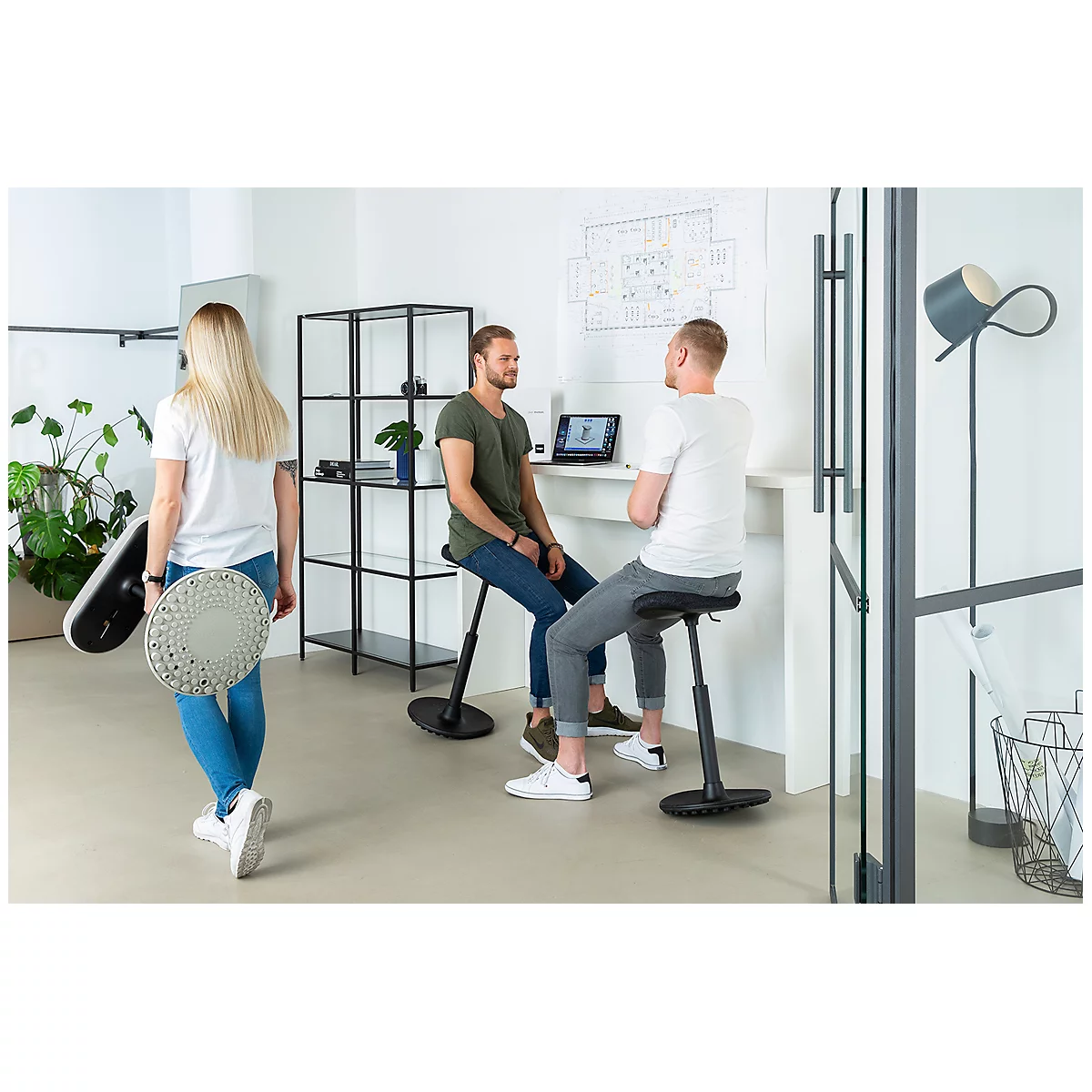 Three people in an office. Two are sitting on ergonomic stools, one person is walking with two stools.