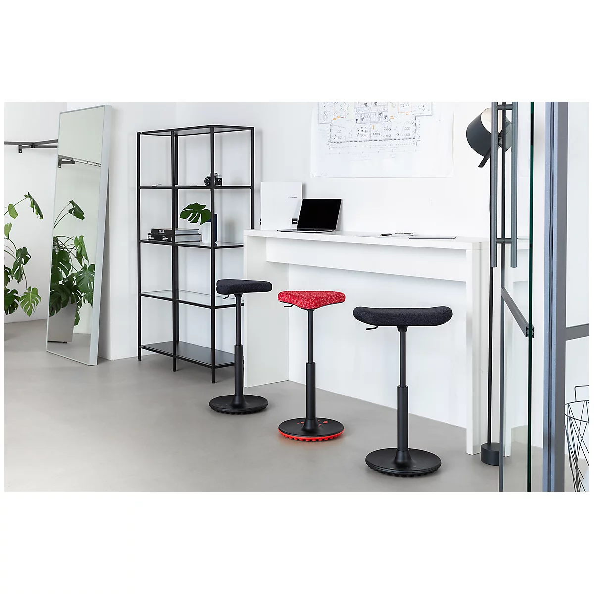 Three standing stools in an office. A black one, a red one, and another black stool stand in front of a white counter. A mirror and a shelf are also visible.