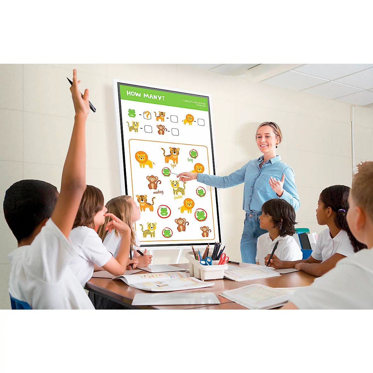 A teacher points to a whiteboard with a counting game. Children in class raise their hands.