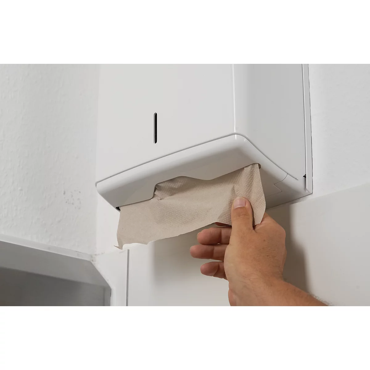 Hand pulling a paper towel from white dispenser on white wall.
