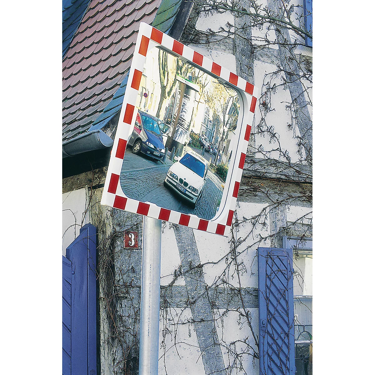 Traffic mirror, red and white framed, shows a street with cars. In the background a half-timbered house with climbing plants. Number 3.