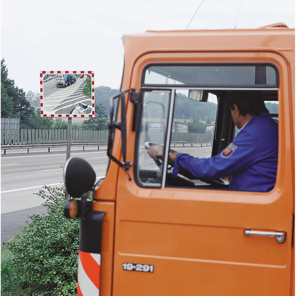 A man driving an orange truck. A convex road traffic mirror is visible through the windshield.