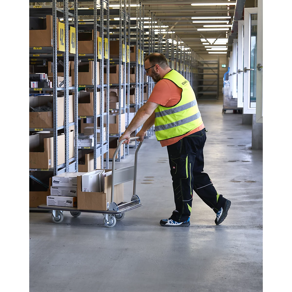 A man in a warehouse pushes a loaded trolley. He wears a yellow safety vest and stands in front of warehouse shelves.