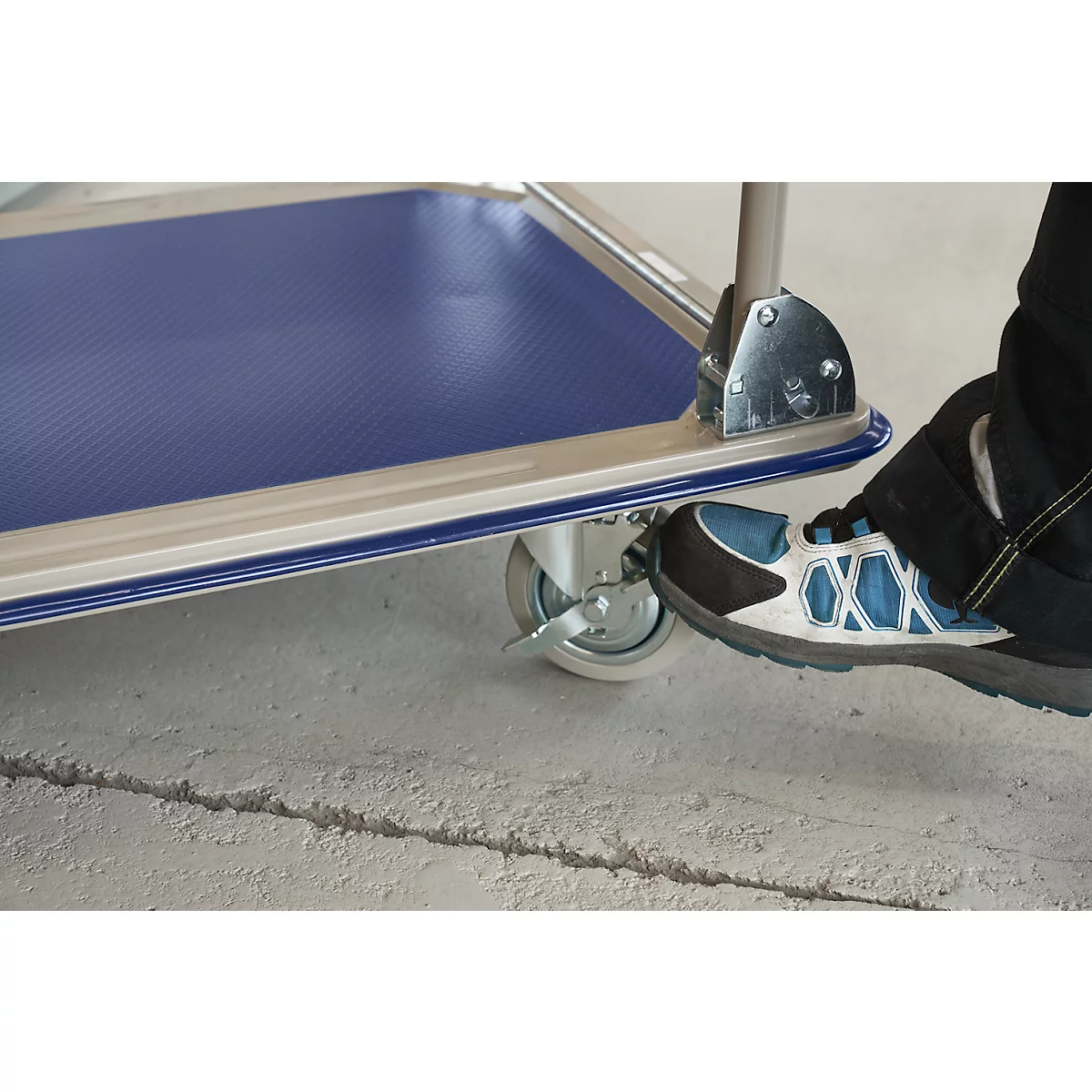 Close-up of a foot activating the parking brake of a trolley; blue loading area, grey ground.