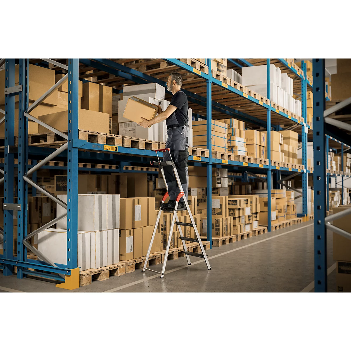 Een man staat op een trap en tilt een doos van een plank in een magazijn.