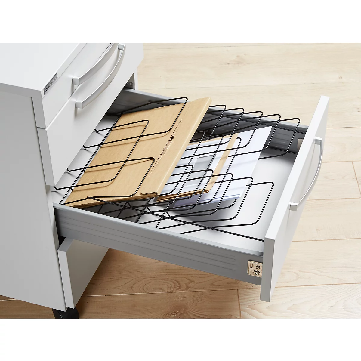 Un tiroir ouvert avec une grille en métal noir et des documents, une armoire gris clair sur un plancher en bois.