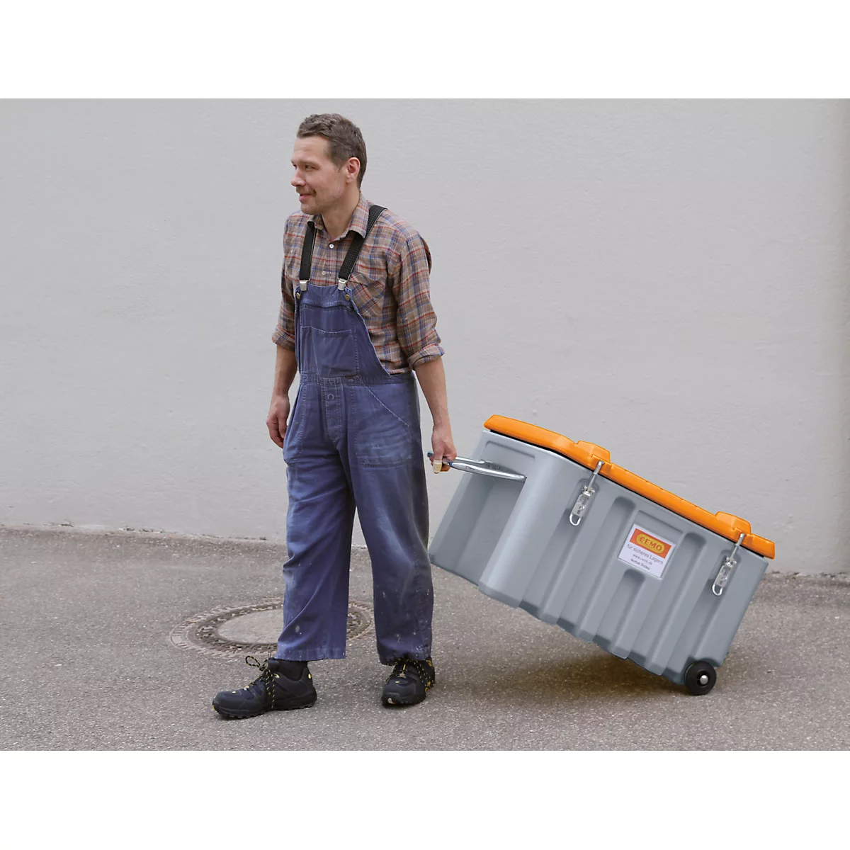 A man in blue overalls pulls a gray suitcase with an orange lid. The suitcase has wheels and a retractable handle.