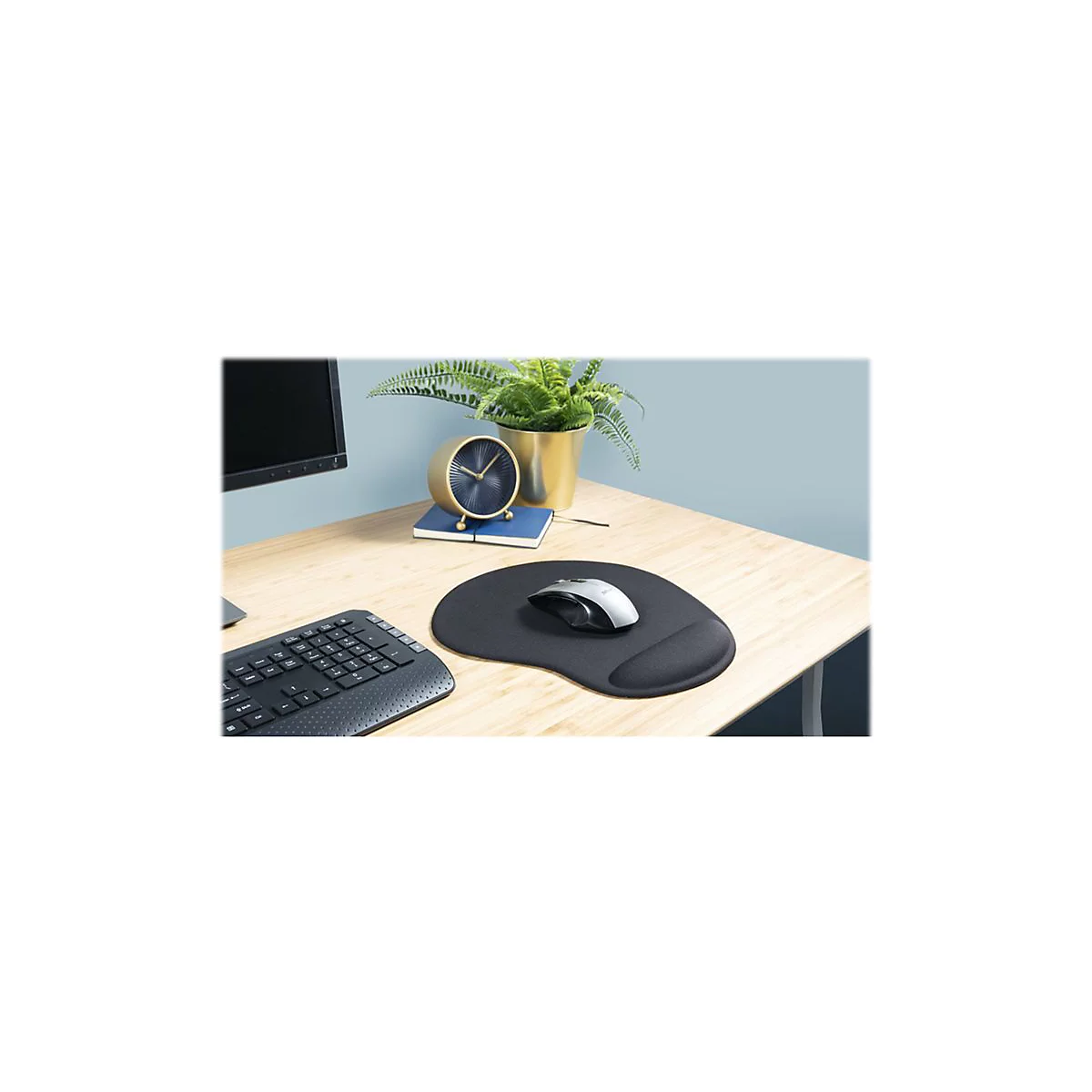 Schwarzes Mauspad mit Handballenauflage, Maus und Tastatur auf Holzschreibtisch. Im Hintergrund eine Uhr und Topf mit Pflanze.