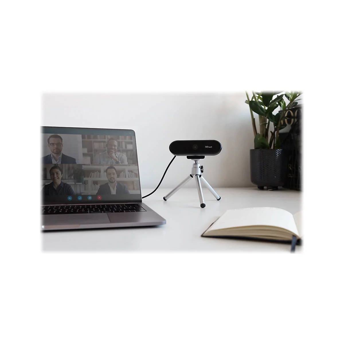 Schwarze Webcam auf Stativ vor Laptop mit Video-Konferenz, neben Pflanze und Notizbuch. Webcam mit Logo Trust.