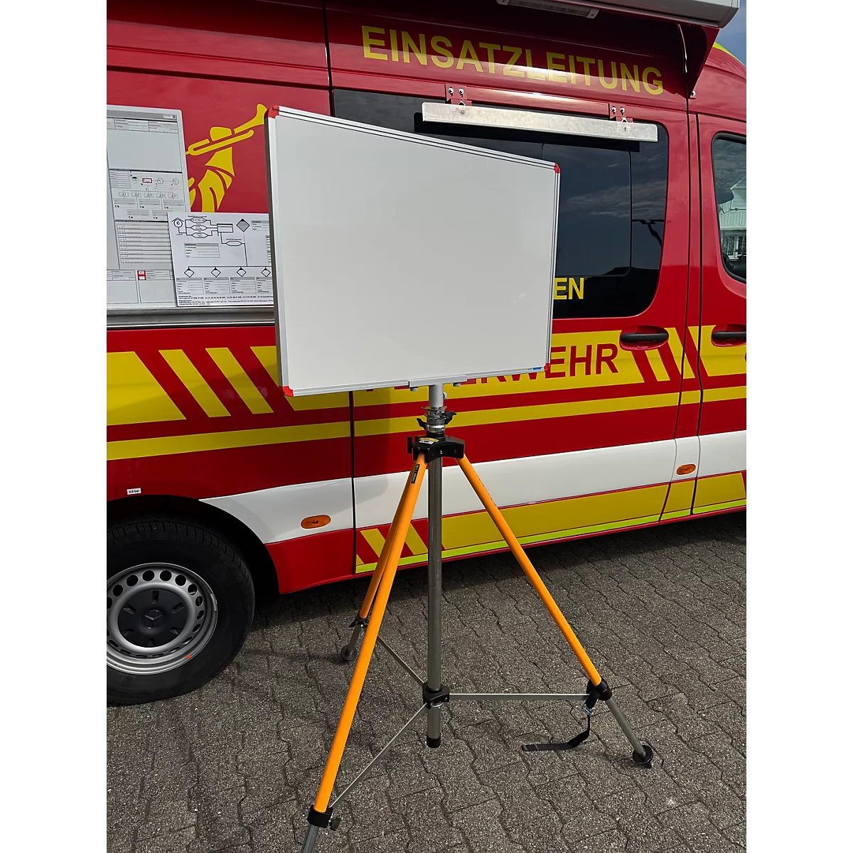 Ein leeres Whiteboard auf einem orangefarbenen Dreibeinstativ vor einem rot-gelben Einsatzleitfahrzeug der Feuerwehr.