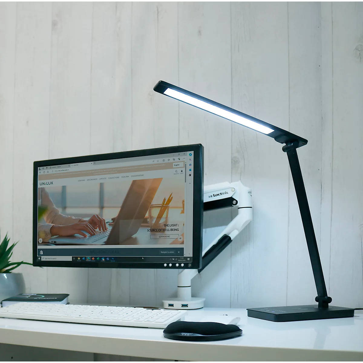 Bureau avec ordinateur et lampe de bureau LED noire allumée. Un site web est visible sur le moniteur.
