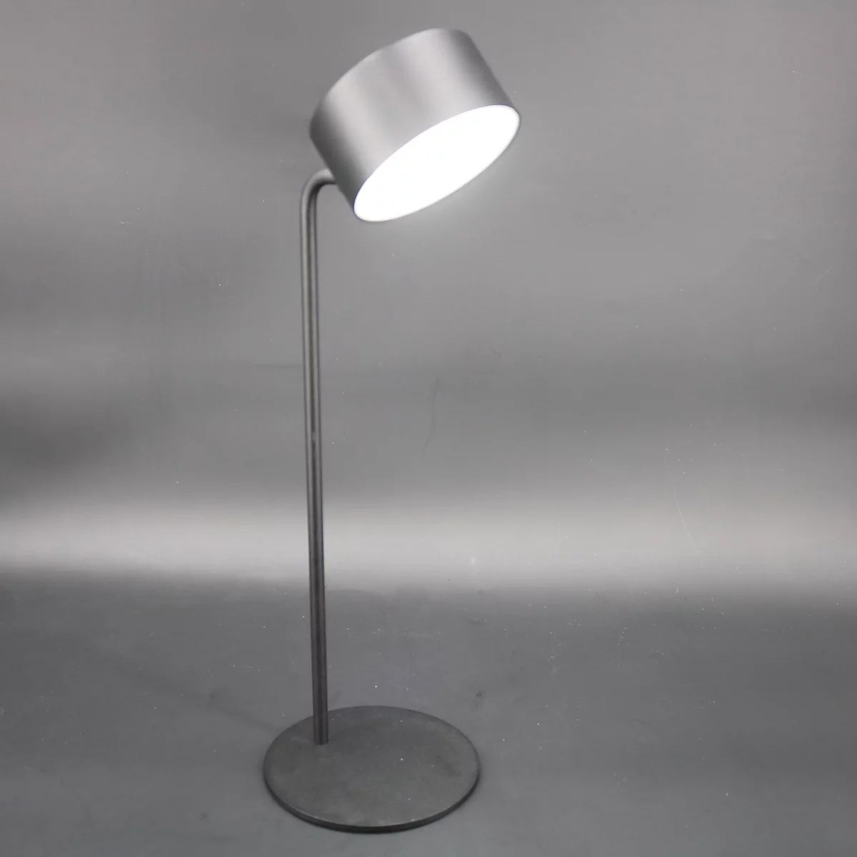 Grijze staande lamp met een cilindrische lampenkap die naar boven is gekanteld. Het licht schijnt fel.