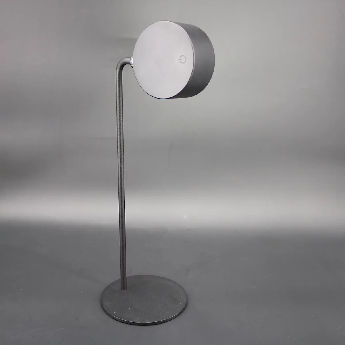 Een metalen vloerlamp. De ronde kop is bevestigd aan een gebogen arm die op een dunne paal staat. Er zit een knop op de kop.