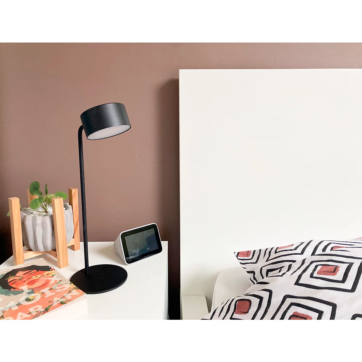 Zwarte tafellamp op nachtkastje naast wit bed, bruine muur. Smart display, plant en boek op tafel.