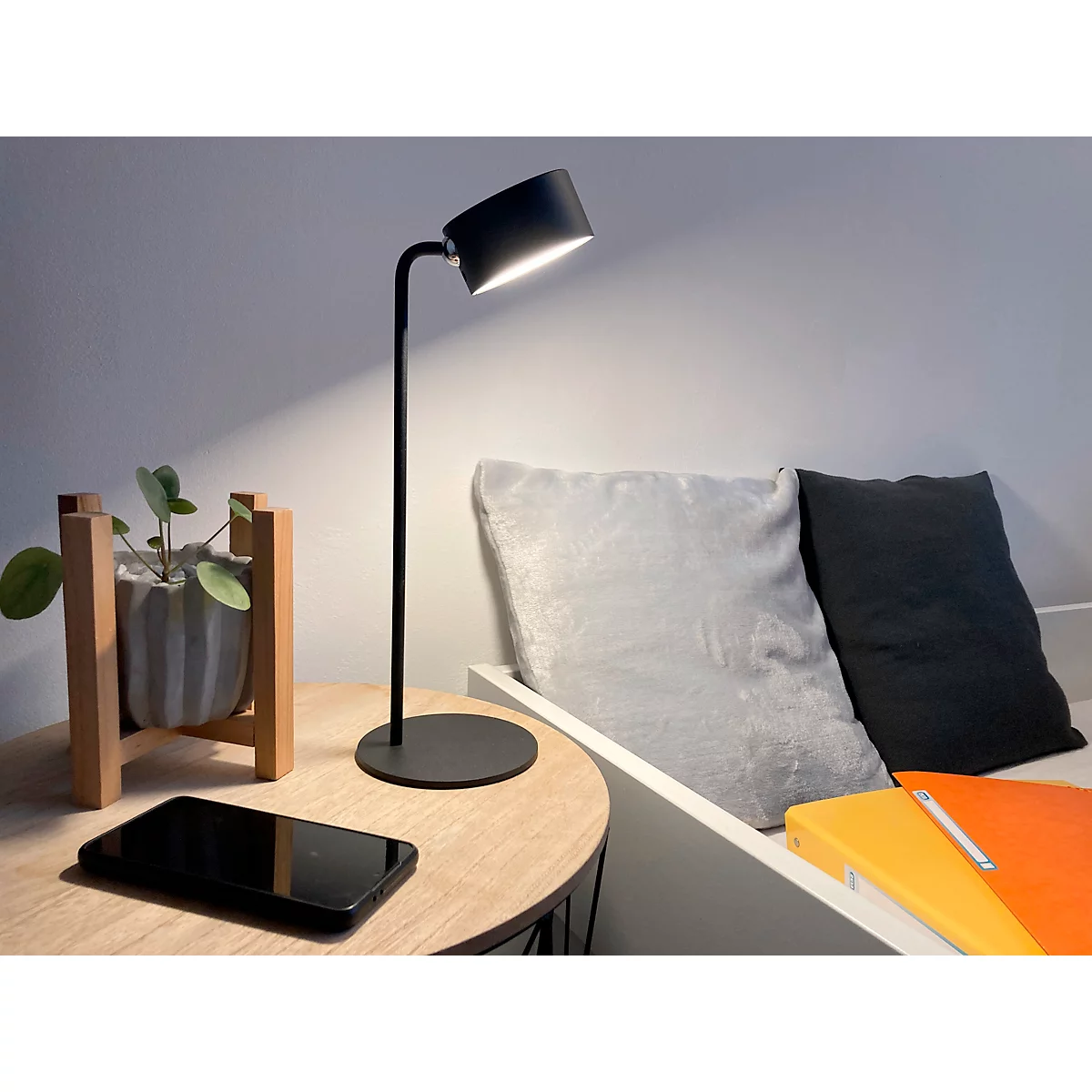 Eine schwarze Tischlampe beleuchtet eine Nachttischszene. Auf dem Tisch liegen ein Smartphone und eine Pflanze.