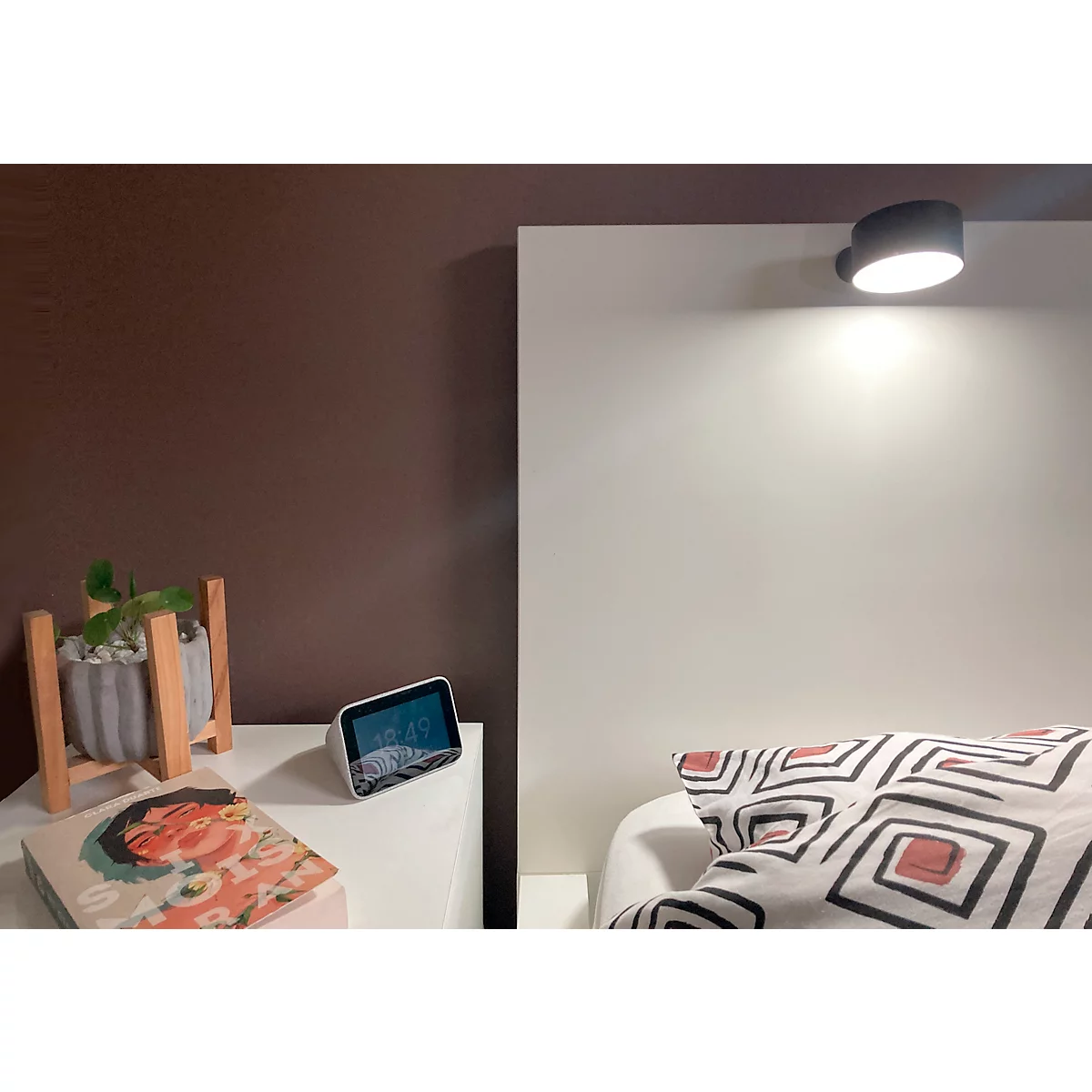 Schlafzimmer mit Nachttisch, Lampe und Kissen. Auf dem Tisch: Buch, Topf mit Pflanze, Wecker.