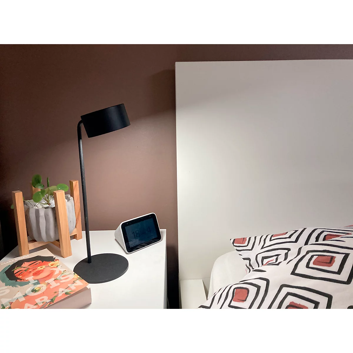 Nachttisch mit Lampe, Pflanze und Smart-Display. Links daneben ein Bett mit Kissen.
