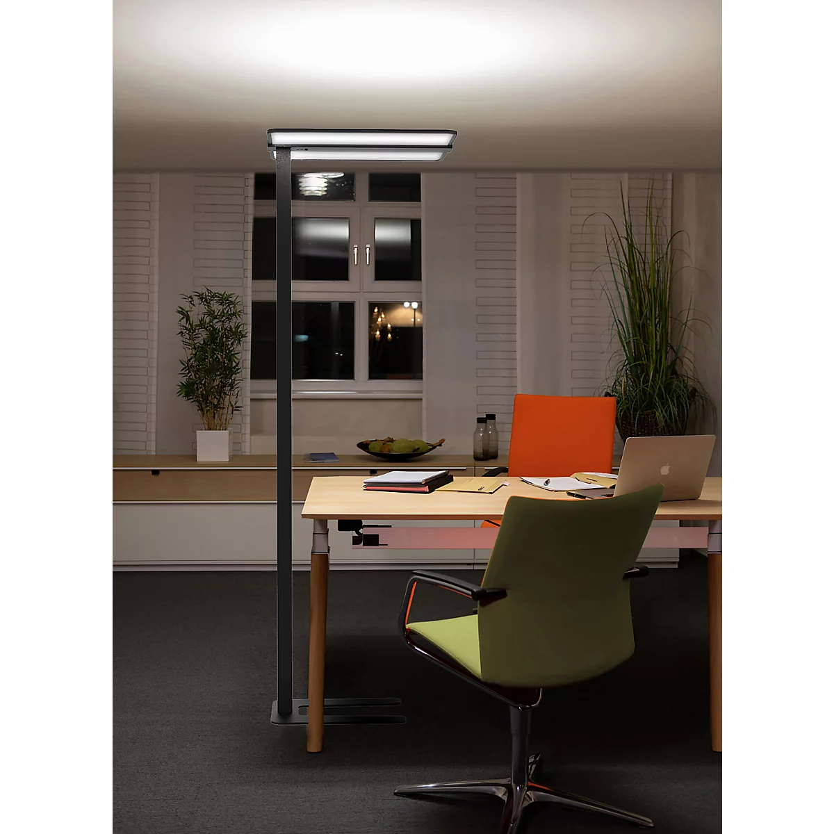 Moderne vloerlamp verlicht een bureau met laptop voor een raam in het donker.
