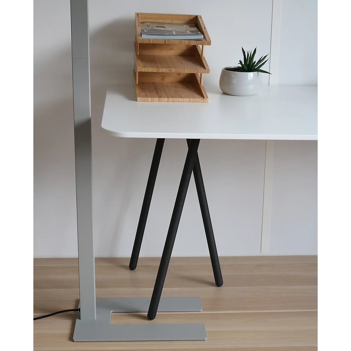 Ein weißer Schreibtisch mit schwarzen Beinen, darauf eine Bambusablage und eine kleine Pflanze vor weißer Wand.