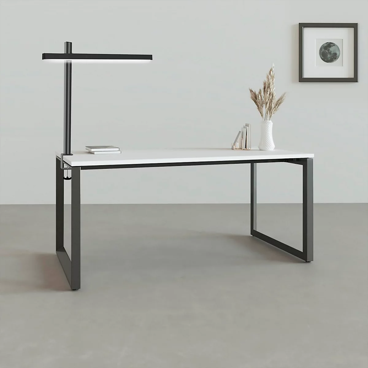 Bureau met zwart frame en wit blad. Boven een zwarte lamp. Op de tafel: boek, vaas met graan. Afbeelding met maan.