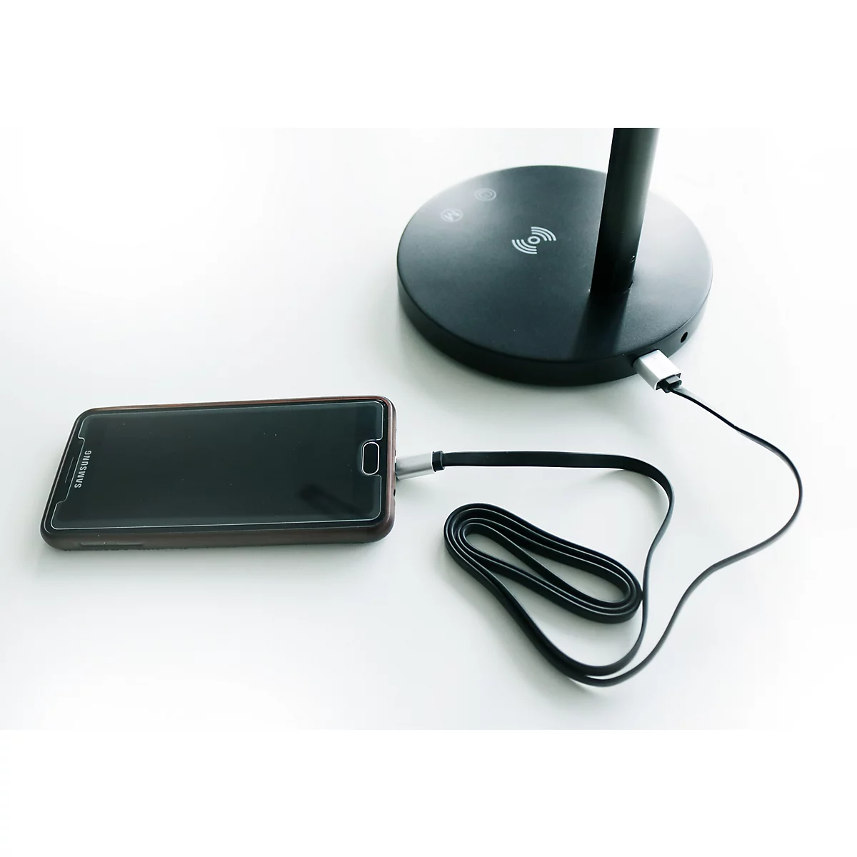 Ein schwarzes Smartphone lädt per Kabel an einem schwarzen Standfuß einer Tischleuchte.