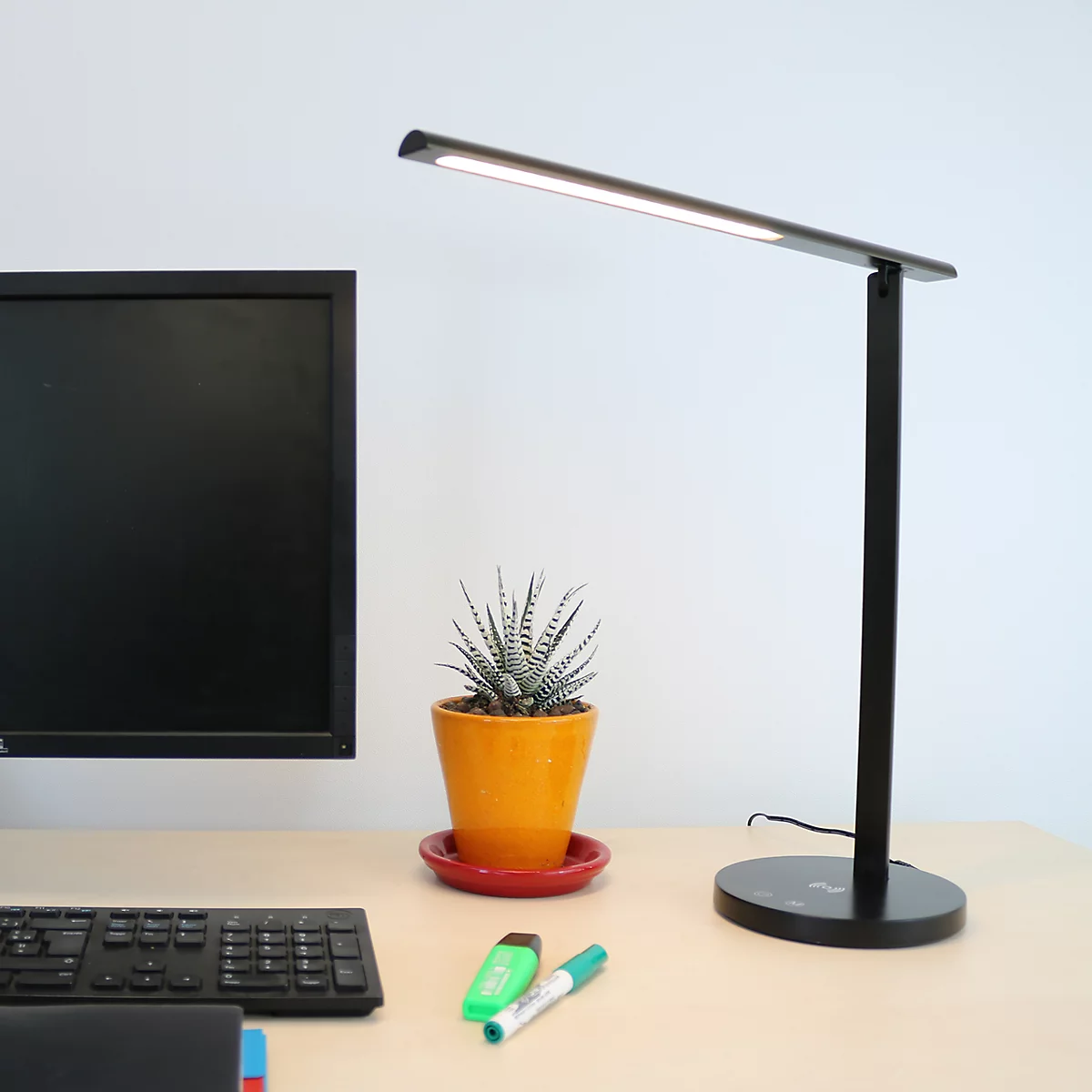 Moderne, schwarze LED-Schreibtischleuchte auf einem Bürotisch neben einem Monitor und einer Topfpflanze.