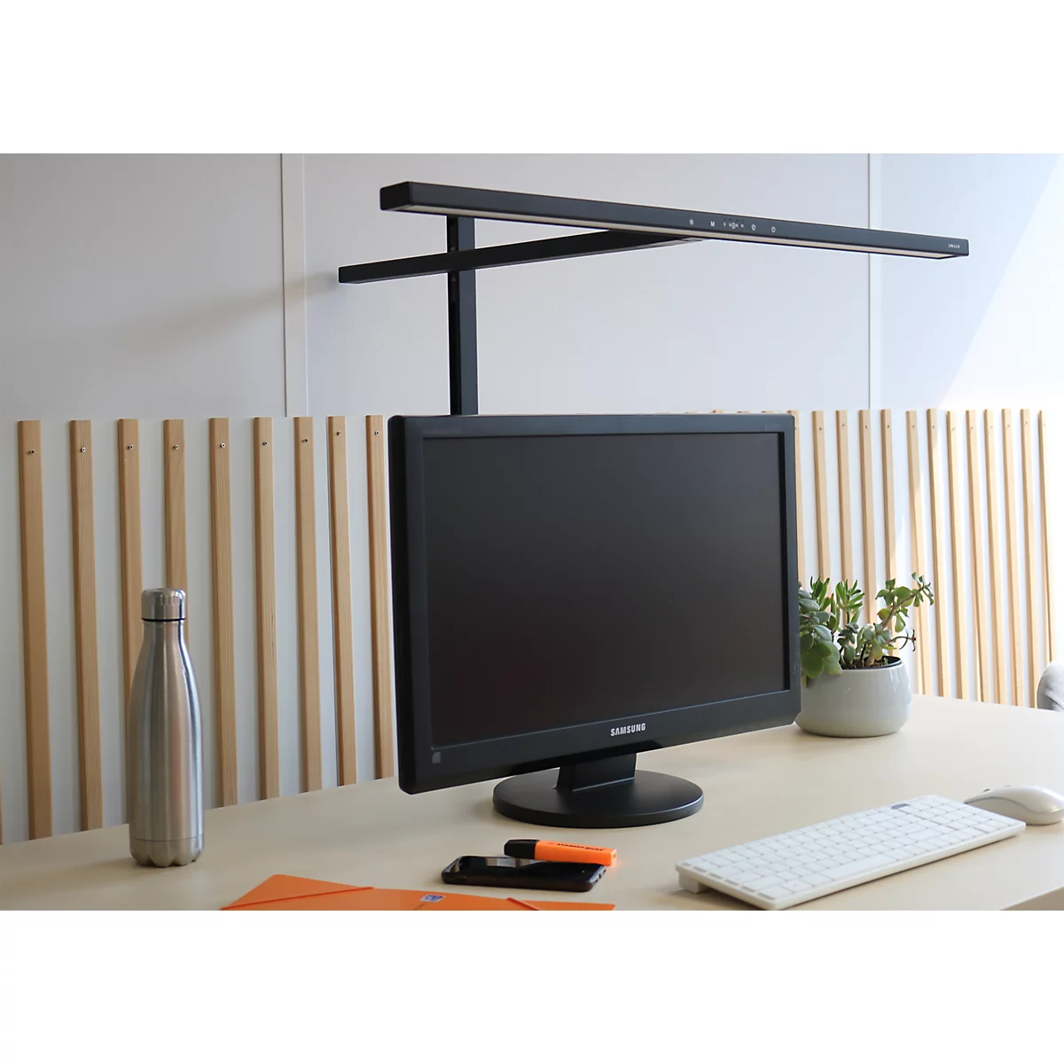Schwarze Monitor-Lichtleiste auf einem hellen Schreibtisch mit Monitor, Tastatur und einer Metall-Trinkflasche.