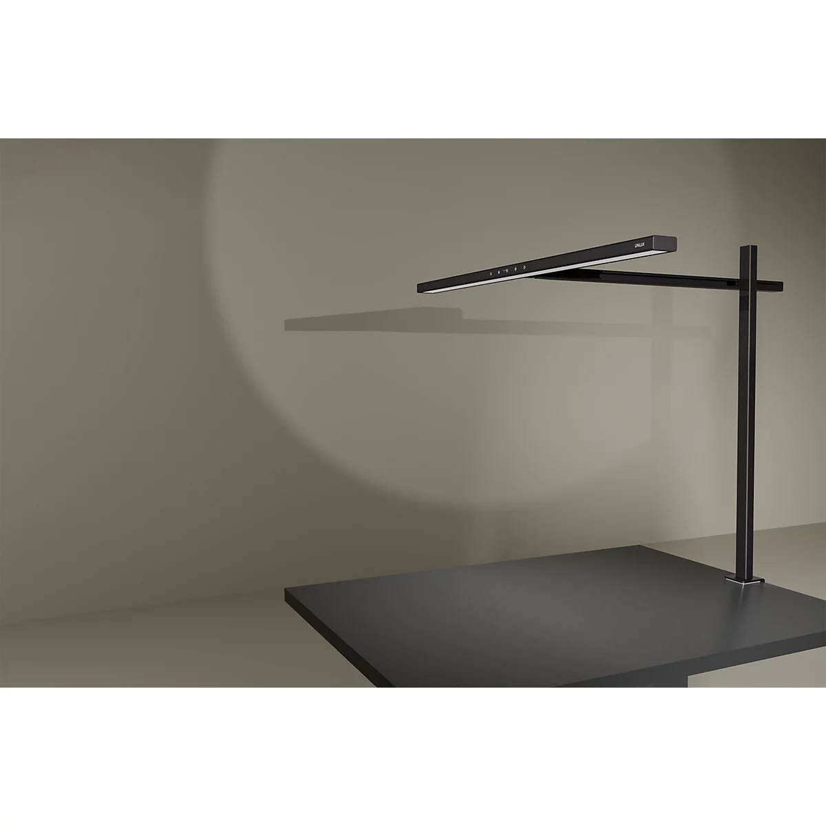 Schwarze, minimalistische Schreibtischlampe leuchtet auf eine graue Wandfläche.