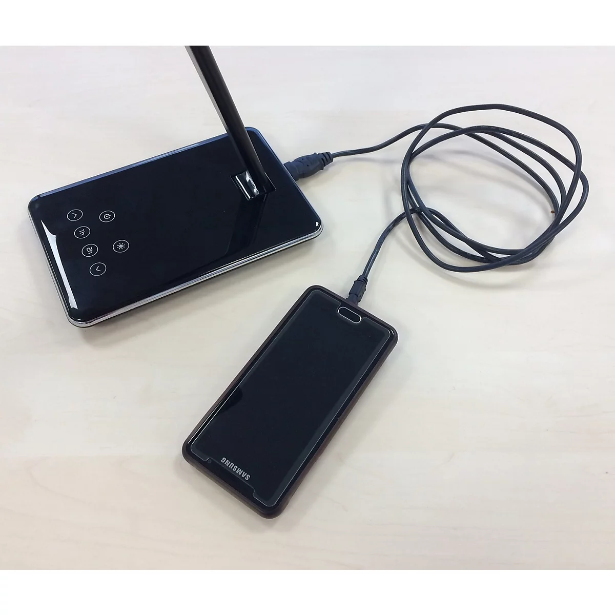 Ein schwarzes Smartphone lädt per USB-Kabel an einer schwarzen LED-Tischleuchte auf hellem Untergrund.