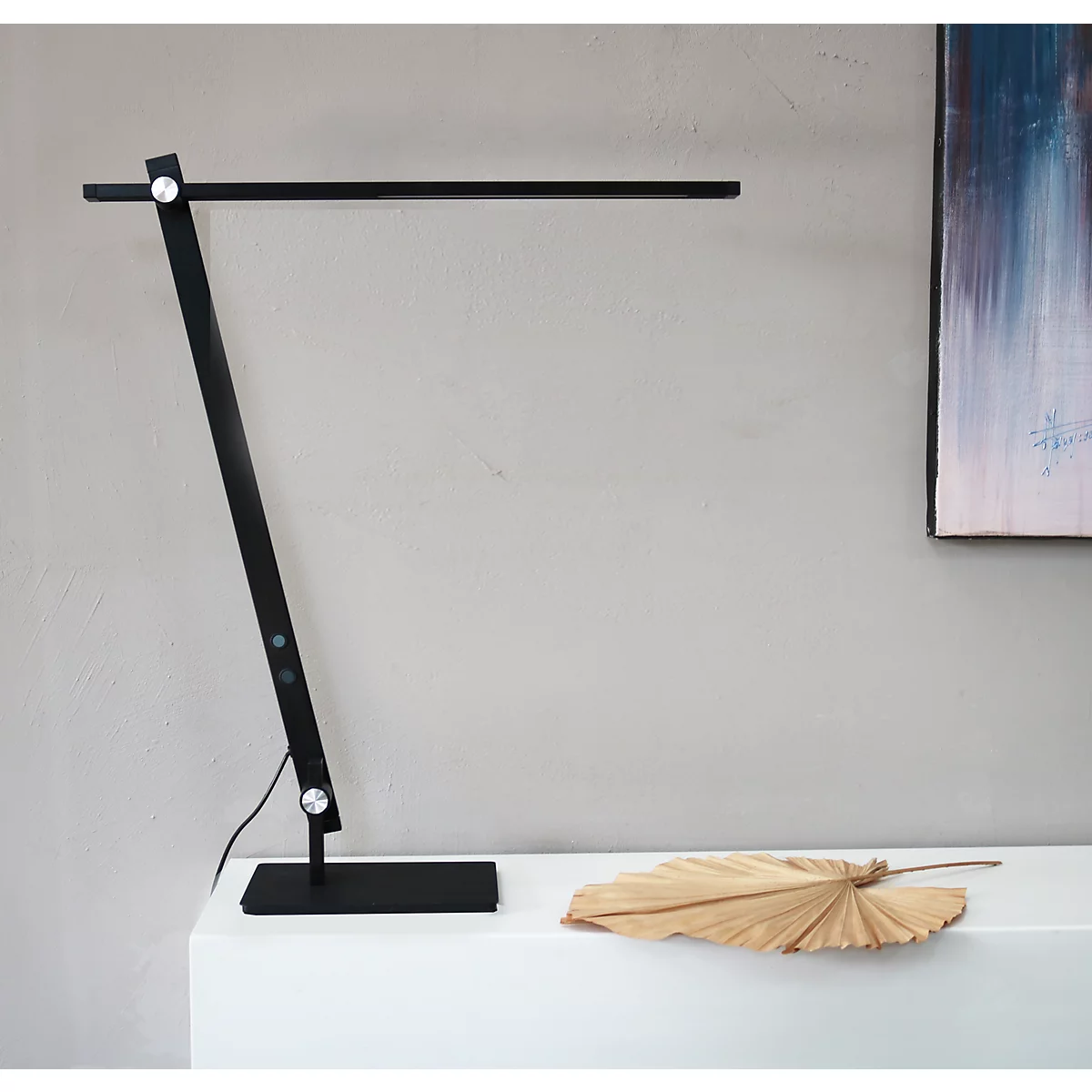 Schwarze, minimalistische Schreibtischleuchte mit schlankem Design auf weißer Ablage neben einem getrockneten Blatt.