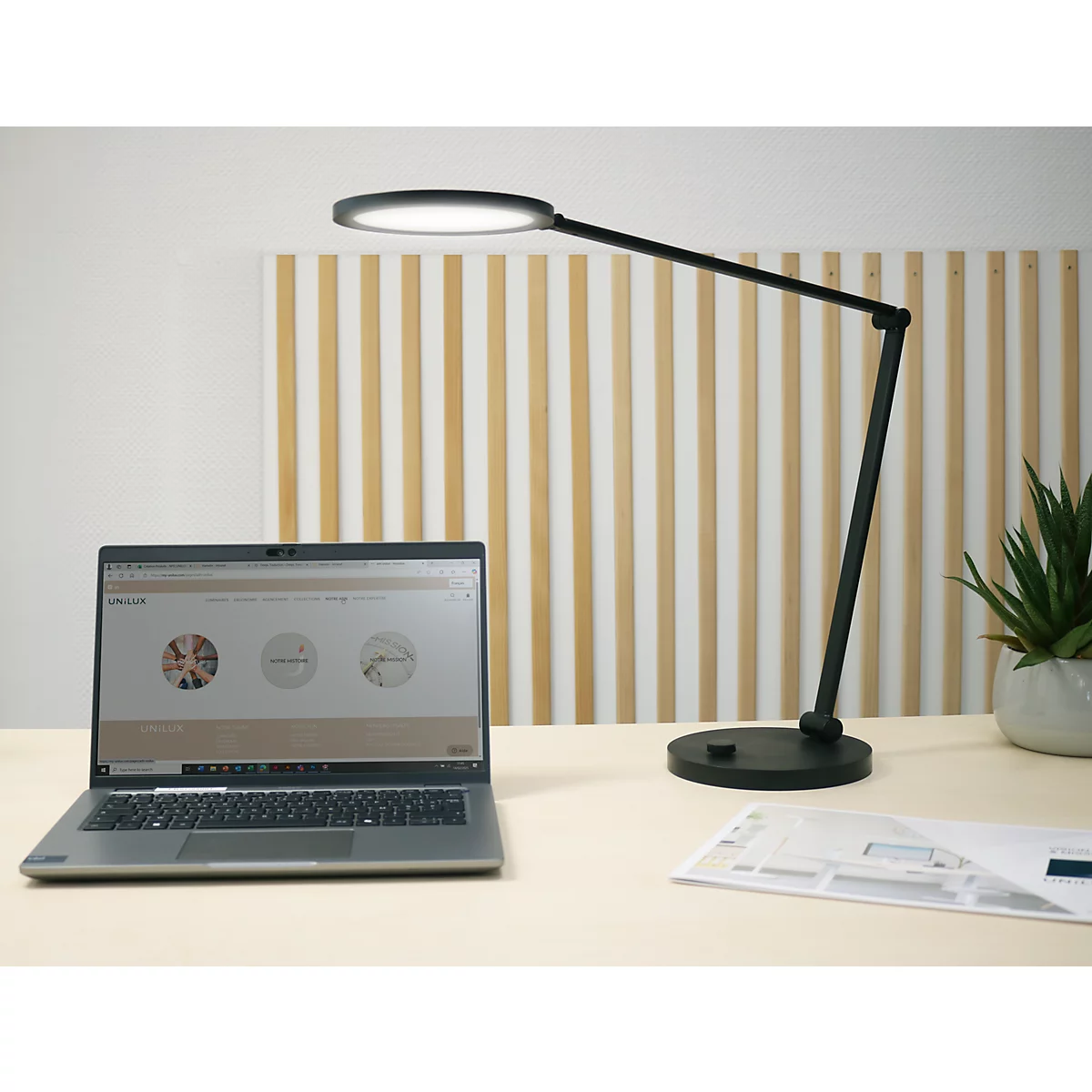 Eine moderne schwarze Schreibtischlampe leuchtet neben einem Laptop auf einem hellen Holztisch.