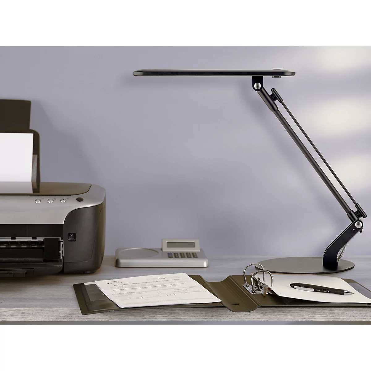 Schwarze Schreibtischlampe leuchtet auf Arbeitsplatz mit Drucker, Taschenrechner und Aktenordner.