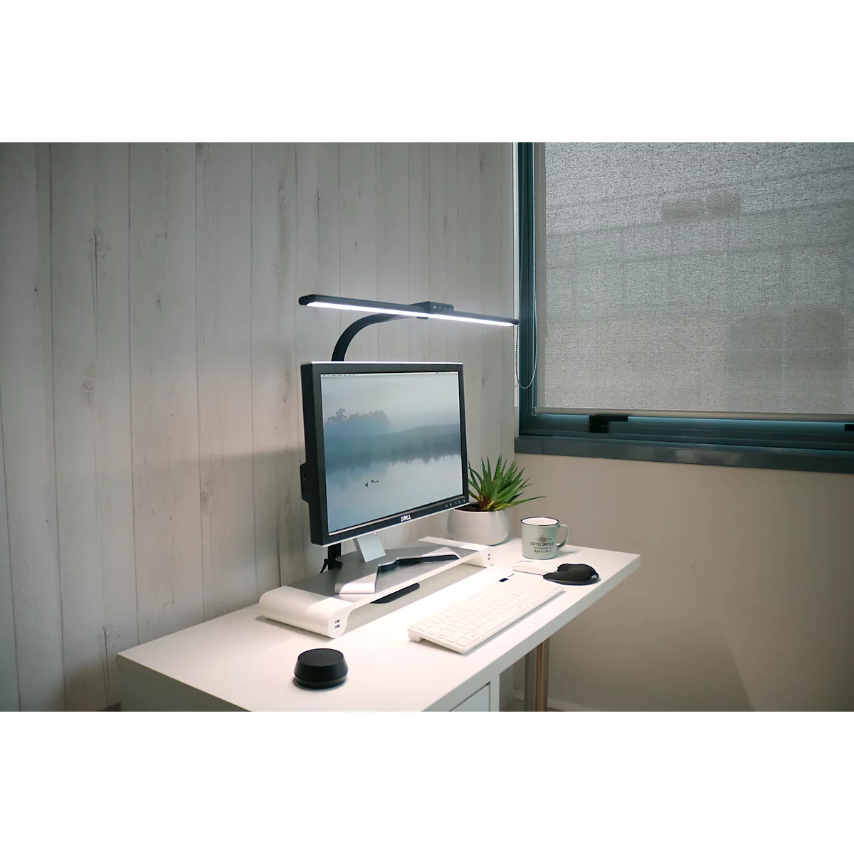 Schreibtisch mit Computer, Lampe und Dekoration. Der Monitor zeigt eine Landschaft, rechts ein Fenster.