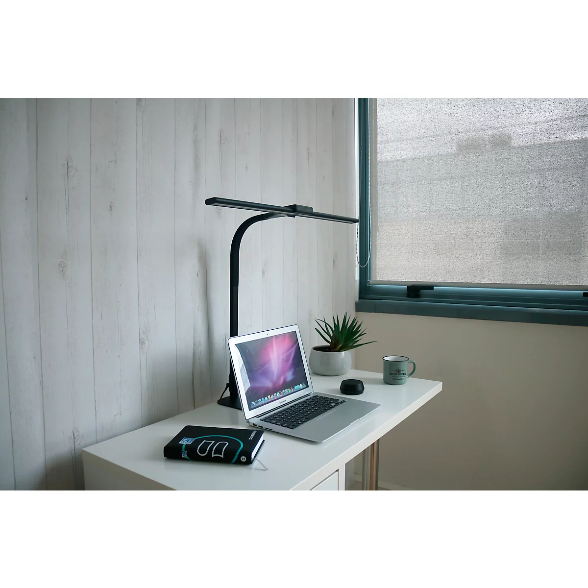 Schreibtisch mit Laptop, Lampe, Pflanze und Becher. Fenster im Hintergrund, helle Holzoptik Wand.
