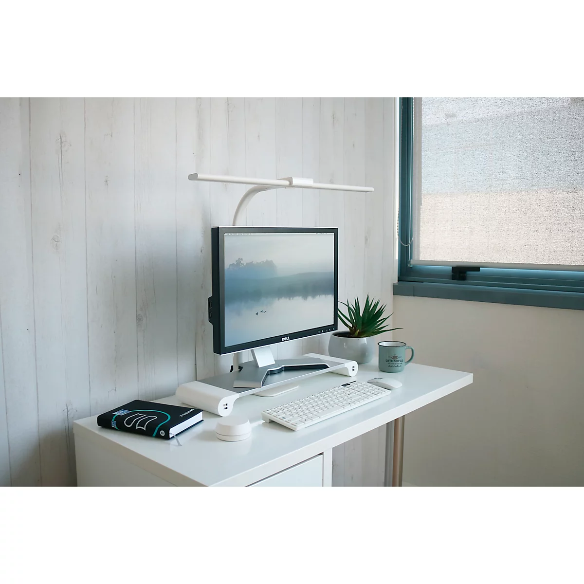 Schreibtisch mit Monitor, Tastatur, Lampe, Pflanze und Fenster. Auf dem Monitor ist eine Naturlandschaft zu sehen.