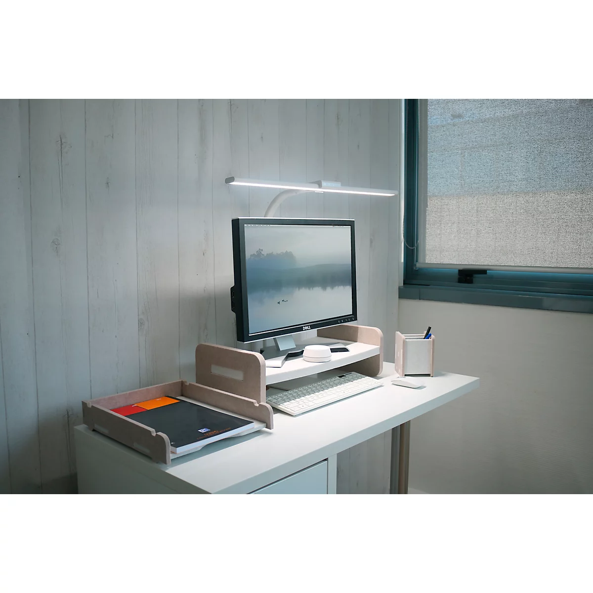 Schreibtisch mit Monitor, Lampe, Ablage und Stiftehalter. Im Hintergrund ein Fenster.